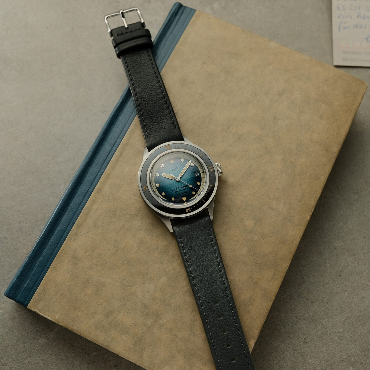 1956 Marine Automatic, stål / blå farveovergang – vintage-inspireret ur fra AV86 Watch med læderrem og blå urskive med farveovergang på en bogbaggrund