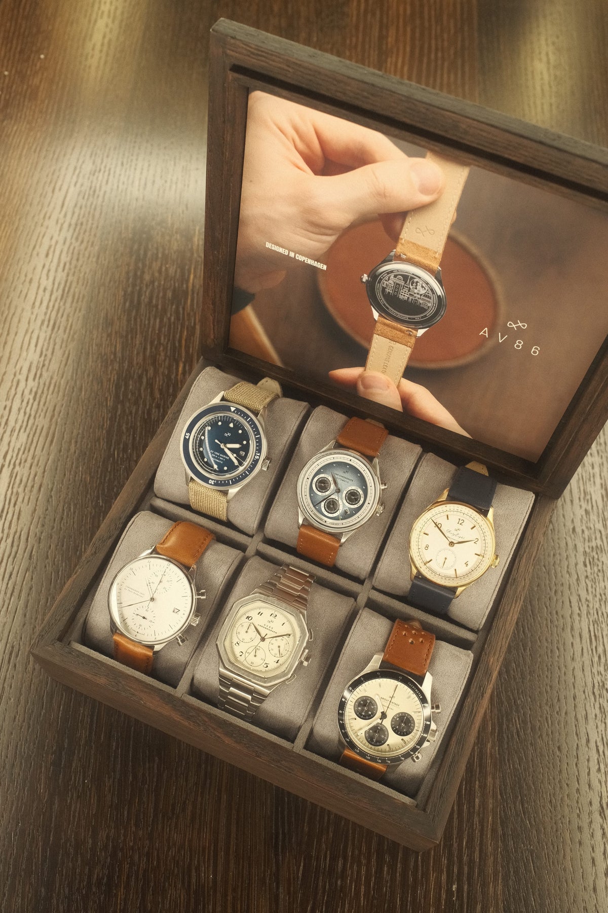 Wooden display box with six diverse wristwatches featuring leather, fabric, and metal straps arranged on soft cushions.