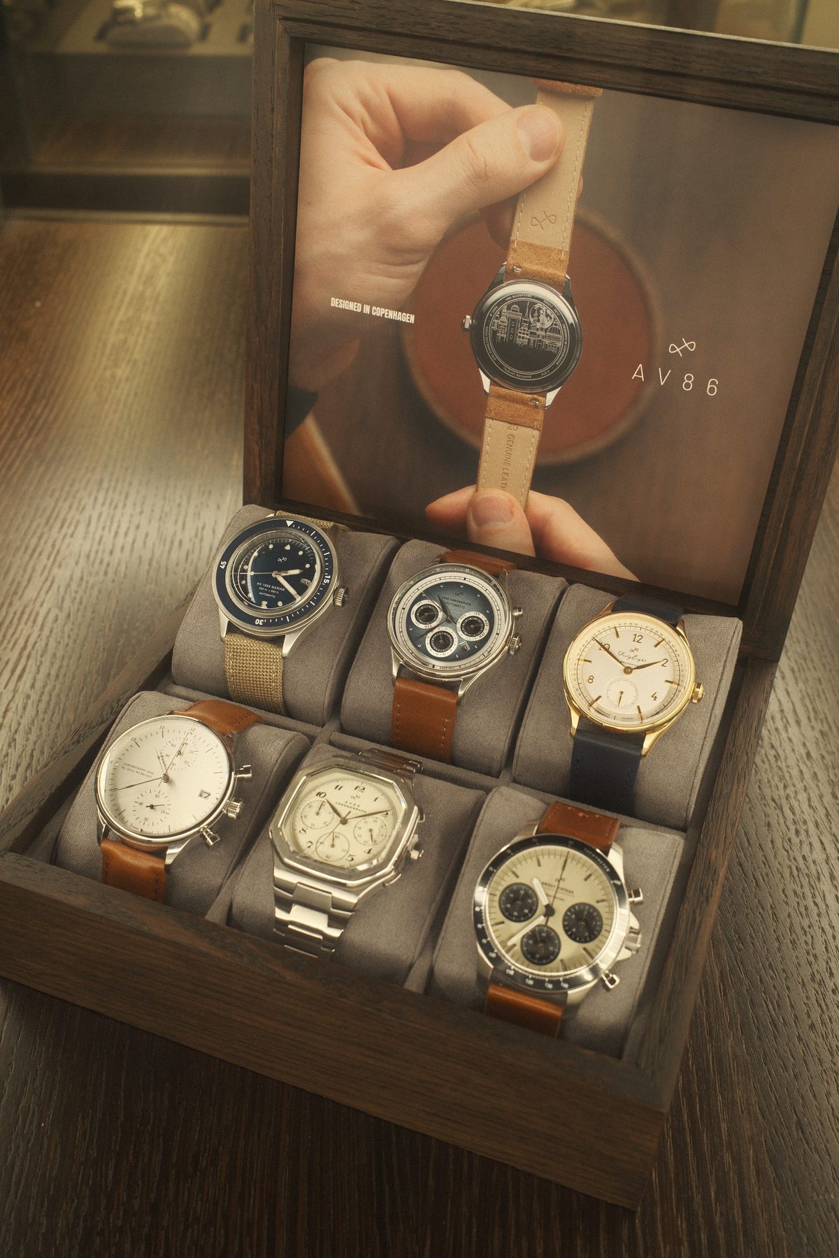 Wooden box with six wristwatches including chronographs, vintage-inspired designs, and leather or metal straps on gray cushions.