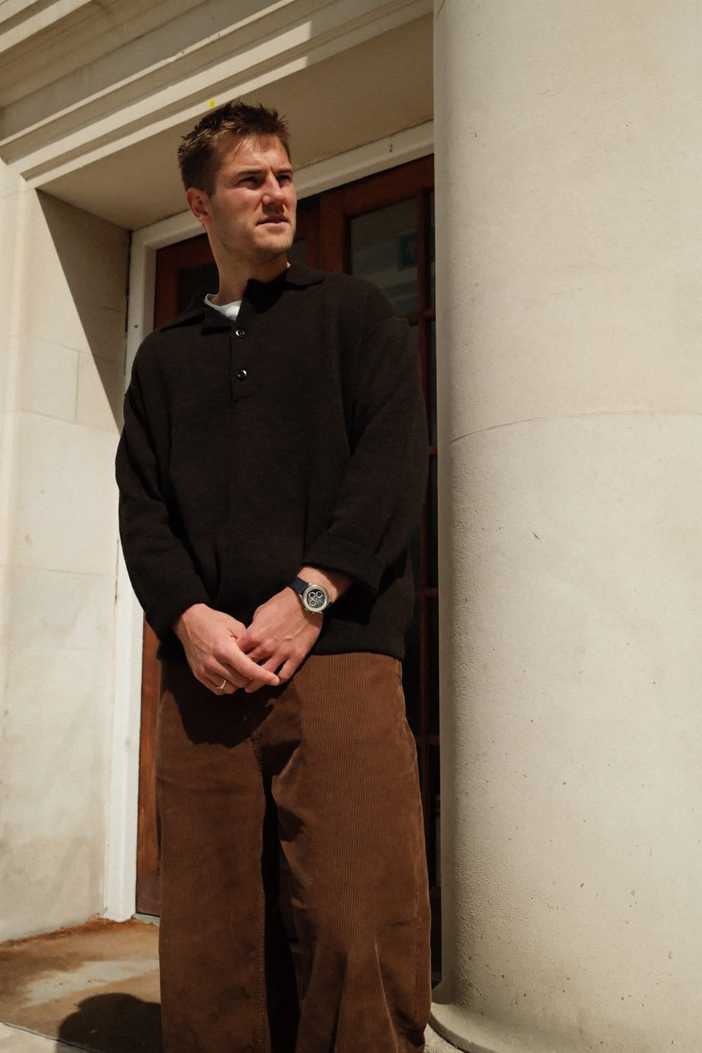 A man stands outside a classical building, dressed in a black knit sweater and brown trousers, wearing the 1564 Chronodate Automatic, Steel / Blue by AV86 on his wrist, capturing a modern Nordic minimalist style with timeless elegance.