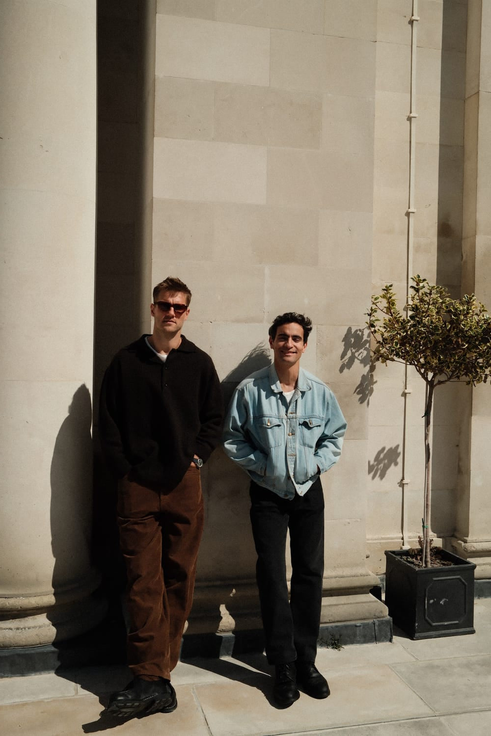 Two men stand casually against a light stone wall in soft sunlight, one wearing brown pants and dark sunglasses, the other in a light denim jacket and black trousers, evoking a relaxed, minimalist Scandinavian style ideal for showcasing the 1564 Chronodate Automatic, Steel / Blue by AV86.