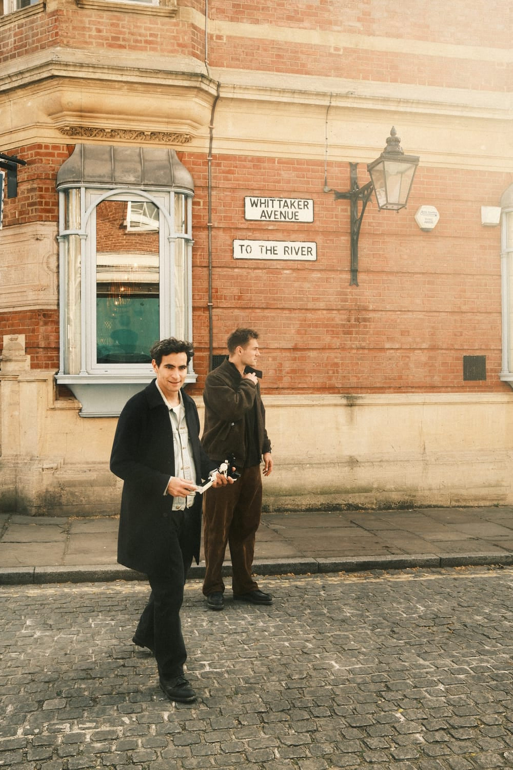 Two men dressed in muted autumn attire walk along a cobblestone street beneath warm sunlight, embodying a relaxed yet sophisticated urban vibe that complements the refined elegance of the 1564 Chronodate Automatic by AV86.