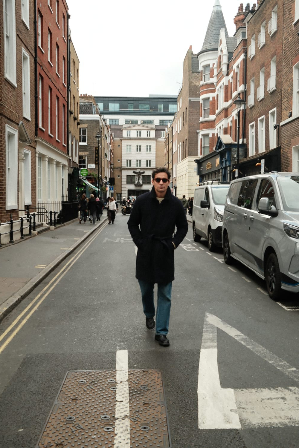 A man wearing sunglasses and a tailored dark coat walks confidently down a city street, his 1564 Chronodate Automatic, Steel / Blue by AV86 subtly visible, embodying understated Scandinavian elegance and urban sophistication.