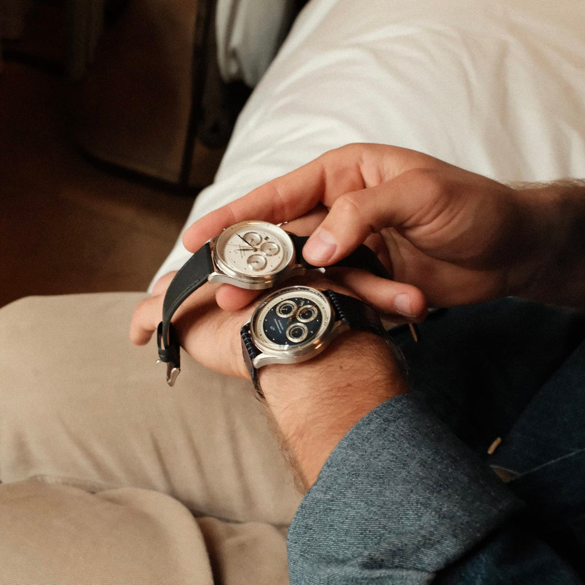 A man wearing a 1564 Chronodate Automatic, Steel / Blue by AV86 on his wrist holds a second vintage-inspired chronograph with a textured dial and leather strap, highlighting refined Danish design and sophisticated horological craftsmanship in a relaxed, intimate setting.