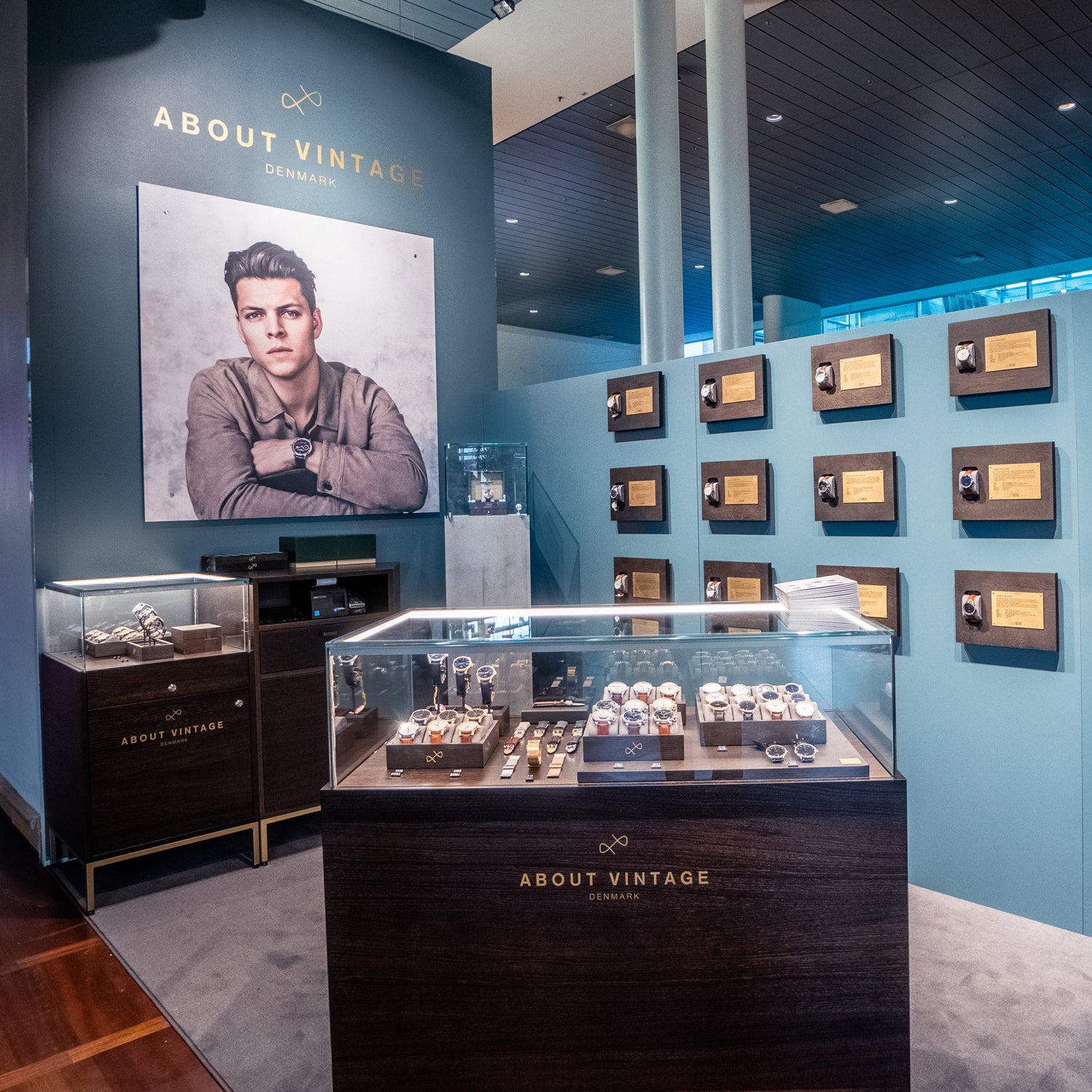 Showcase area featuring vintage-inspired wristwatches in glass cases with a large portrait and award plaques on blue walls background.