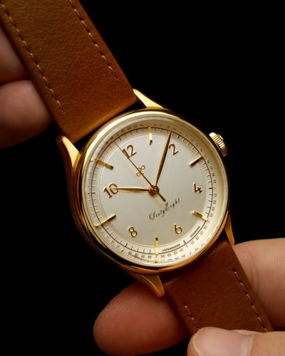 Classic gold-tone watch with clean white dial, Arabic numerals, and brown leather strap, held against a dark background.